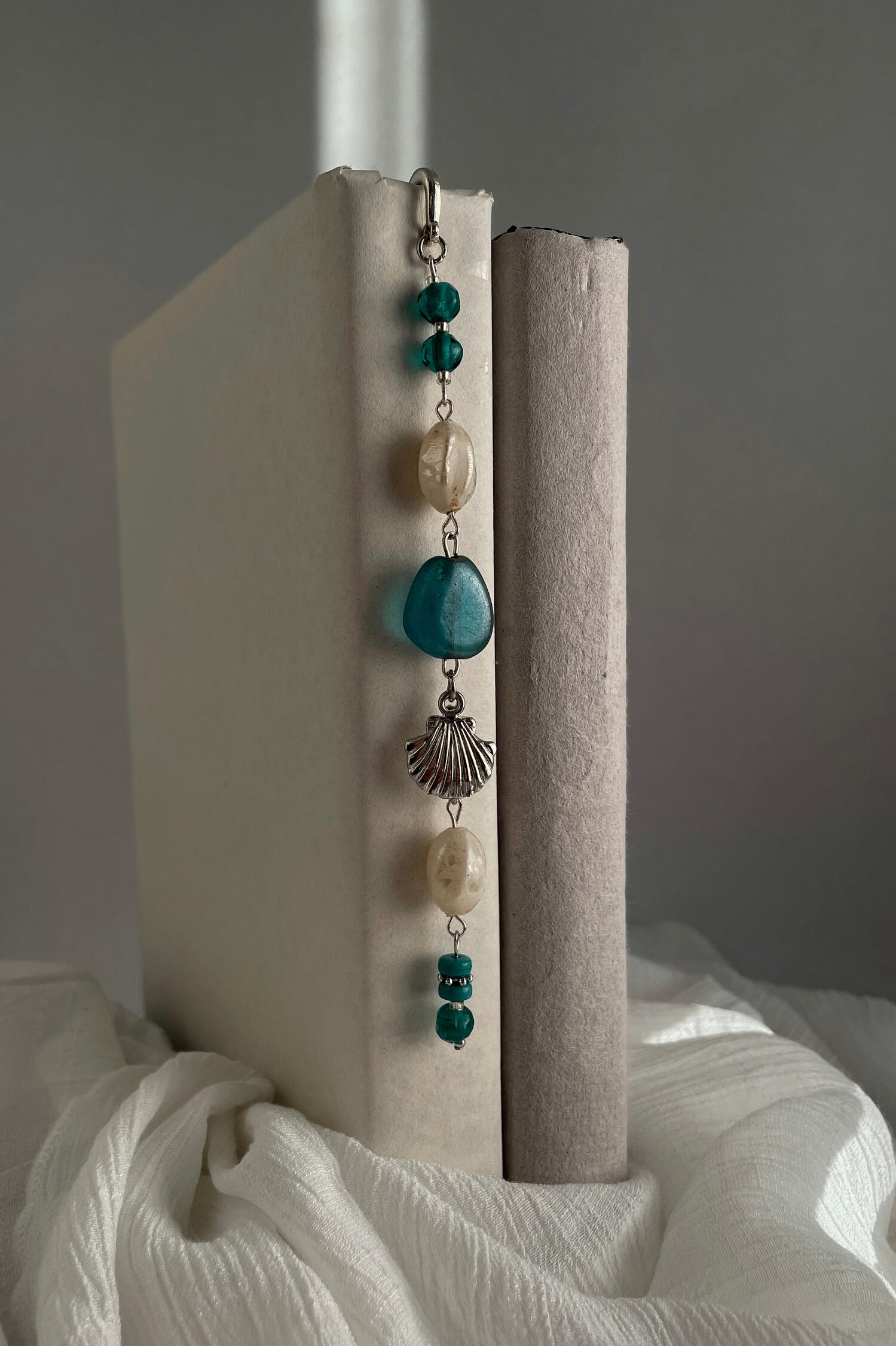 Handmade beaded bookmark featuring silver hardware, turquoise and cream glass beads, and a shell charm, photographed resting between the pages of an open book.